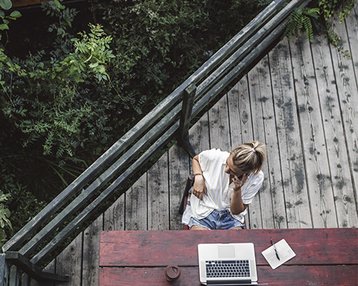 Eine Person sitzt an einem Holztisch im Freien, telefoniert und arbeitet an einem Laptop. Neben dem Laptop liegt ein Notizbuch. Umgeben von Pflanzen und Holzdeck.