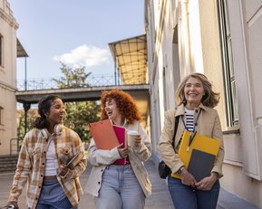 Drei Frauen gehen lächelnd auf einem Gehweg, tragen Ordner und Getränke, umgeben von Gebäuden und Tageslicht.