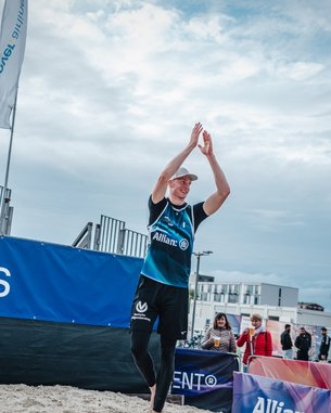 Ein Sportler in einem blauen Trikot applaudiert auf einem Sandplatz, umgeben von Zuschauern und Werbebannern. Die Atmosphäre ist sportlich und die Veranstaltung scheint ein Beachvolleyballspiel zu sein.