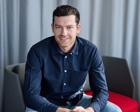 Ein junger Mann mit dunklem Hemd sitzt auf einem Stuhl und lächelt, während er die Hände auf den Knien hält.