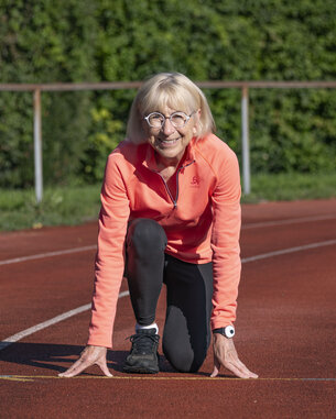 Eine ältere Frau in sportlicher Kleidung kniet auf einer Laufbahn und bereitet sich auf den Start eines Rennens vor. Sie trägt eine orangefarbene Jacke und hat eine sportliche Haltung.