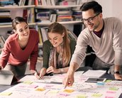 Drei Personen arbeiten an einem Tisch mit Notizen und einem großen Blatt Papier. Sie diskutieren Ideen und schreiben auf, während Bücherregale im Hintergrund sichtbar sind.