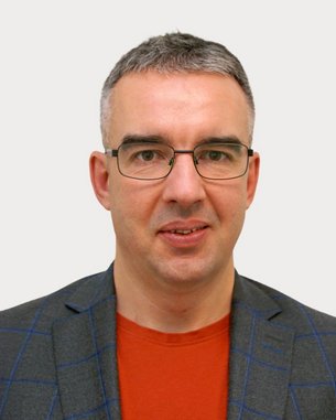 Mann mit Brille, grauen Haaren und einem grauen Blazer mit Karomuster, trägt ein rotes T-Shirt. Neutraler Hintergrund.