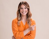 Eine lächelnde Frau mit langen, lockigen Haaren trägt ein orangefarbenes Oberteil und steht mit verschränkten Armen. Der Hintergrund ist neutral und hell.