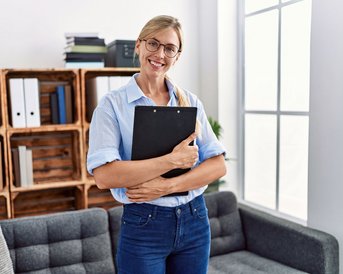 Eine Frau mit Brille steht in einem modernen Raum und hält eine Mappe. Im Hintergrund sind Regale mit Ordnern und Pflanzen zu sehen.