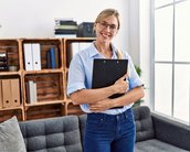 Eine Frau mit Brille steht in einem modernen Raum und hält eine Mappe. Im Hintergrund sind Regale mit Ordnern und Pflanzen zu sehen.