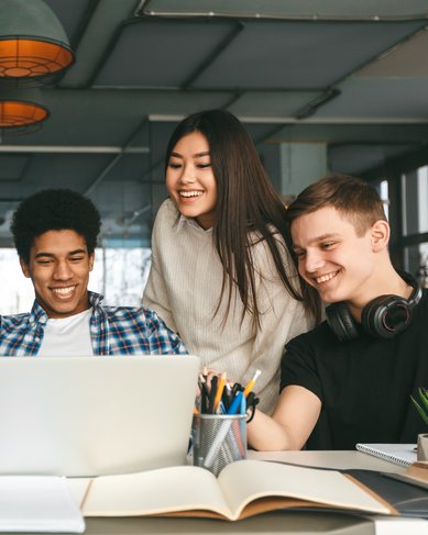 Sechs junge Erwachsene sitzen an einem Tisch und schauen auf einen Laptop. Sie wirken interessiert und lachen, während sie gemeinsam an einem Projekt arbeiten. Bücher und Schreibutensilien sind auf dem Tisch verteilt.