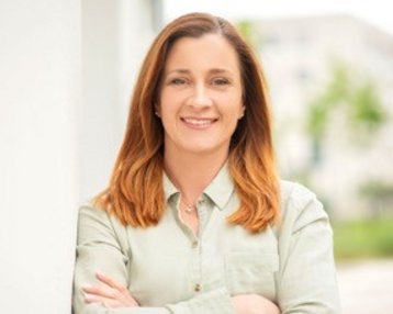 Eine Frau mit langen, braunen Haaren steht lächelnd mit verschränkten Armen vor einem unscharfen Hintergrund. Sie trägt ein helles Hemd und wirkt selbstbewusst.