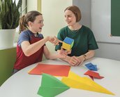 Eine Lehrerin und ein Schüler sitzen an einem Tisch und diskutieren über bunte Papierformen, während sie eine geometrische Figur halten.