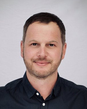 Ein Mann mit kurzem, dunklem Haar und einem leichten Bart lächelt in die Kamera. Er trägt ein dunkelblaues Hemd und hat einen neutralen Gesichtsausdruck vor einem hellen Hintergrund.