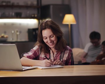 Eine Frau sitzt an einem Tisch, schreibt in ein Notizbuch und lächelt, während ein Laptop vor ihr steht. Im Hintergrund sind zwei Personen zu sehen.