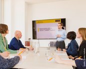 Eine Gruppe von sechs Personen sitzt an einem Tisch und hört einem Referenten zu, der vor einem Bildschirm mit Präsentation steht. Auf dem Bildschirm sind Informationen zu Rechtschreibung und Grammatik zu sehen.