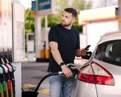 Ein Mann tankt sein Auto an einer Tankstelle und schaut auf sein Smartphone. Mehrere Zapfsäulen sind im Hintergrund sichtbar.