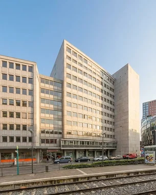 Moderne Bürogebäude mit Glasfassade und asymmetrischer Architektur, umgeben von Straßen und Schienen. Der Himmel ist bewölkt, was eine gedämpfte Lichtstimmung erzeugt.