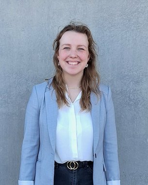 Eine lächelnde Frau steht vor einer grauen Wand. Sie trägt einen hellgrauen Blazer, ein weißes Hemd und schwarze Jeans. Ihre Schuhe sind weiß, und sie hat lange, lockige Haare.