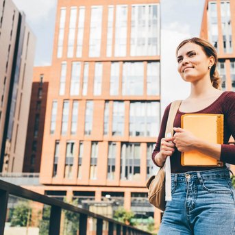 Eine junge Frau mit einem Notizbuch und einer Tasche steht in einem urbanen Umfeld mit modernen Gebäuden im Hintergrund. Sie trägt ein langärmliges Oberteil und Jeans.