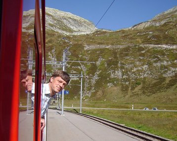 Ein Mann lehnt aus dem Fenster eines Zuges, während er an einem Bahnhof mit einer Bank und Gleisen vorbeifährt. Im Hintergrund sind grüne Hügel und Felsen sichtbar.