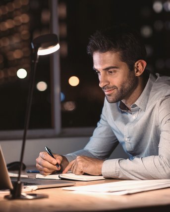 Ein Mann sitzt an einem Schreibtisch, schreibt in ein Notizbuch und arbeitet am Laptop. Er ist von einer Schreibtischlampe beleuchtet, während die Stadtlichter im Hintergrund sichtbar sind.