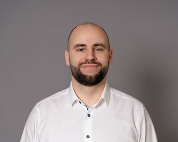 Ein Mann mit kurzem Haar und Bart trägt ein weißes Hemd. Er steht vor einem grauen Hintergrund und lächelt leicht.