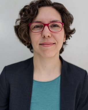 Eine Person mit lockigem, braunem Haar und roten Brillen sitzt vor einem hellen Hintergrund. Sie trägt ein dunkelblaues Blazer und ein türkisfarbenes Oberteil.