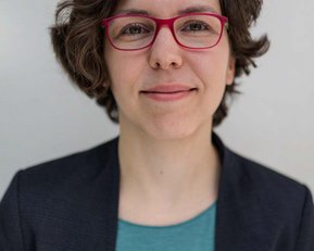 Eine Person mit lockigem, braunem Haar und roten Brillen sitzt vor einem hellen Hintergrund. Sie trägt ein dunkelblaues Blazer und ein türkisfarbenes Oberteil.