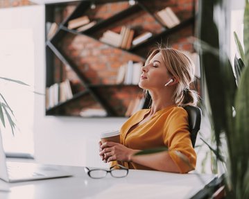 Resilienz Übungen Eine Frau mit Kopfhörern sitzt entspannt an einem Schreibtisch, hält einen Kaffeebecher in der Hand und blickt nach oben. Im Hintergrund sind Bücherregale und Pflanzen sichtbar.