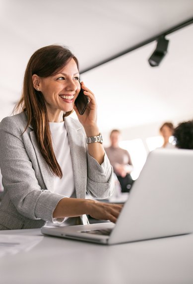 Eine Frau sitzt am Schreibtisch, spricht am Telefon und tippt auf einem Laptop. Im Hintergrund sind weitere Personen in einem modernen Büro zu sehen.