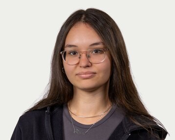 Junge Frau mit langen, glatten Haaren und Brille, trägt einen schwarzen Hoodie und eine graue Bluse. Sie schaut direkt in die Kamera.