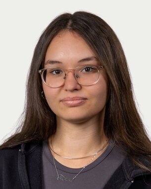 Junge Frau mit langen, glatten Haaren und Brille, trägt einen schwarzen Hoodie und eine graue Bluse. Sie schaut direkt in die Kamera.