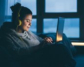 Eine Person sitzt auf einem Sofa, trägt Kopfhörer und schaut auf einen Laptop. Die Umgebung ist schwach beleuchtet, und es sind Fenster im Hintergrund zu sehen.