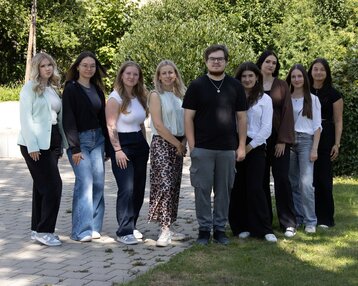 Eine Gruppe von neun Personen steht auf einem Gehweg, umgeben von Bäumen und Grünflächen, in lässiger Kleidung.