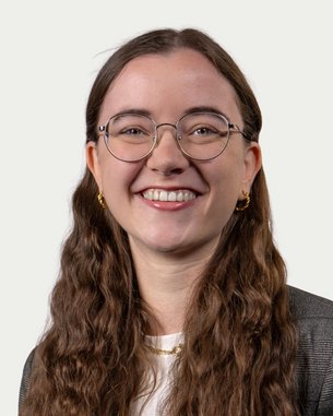 Eine lächelnde Frau mit langen, lockigen Haaren und Brille trägt einen Blazer. Der Hintergrund ist neutral und hell.