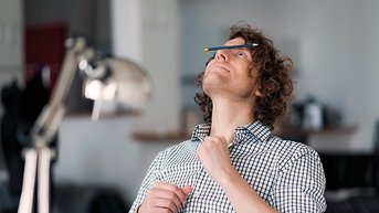 Ein Mann mit lockigem Haar sitzt an einem Tisch, während er einen Stift auf seiner Stirn balanciert. Er trägt ein kariertes Hemd und schaut nach oben.