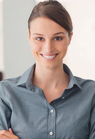 Eine lächelnde Frau in einem blauen Hemd steht mit verschränkten Armen in einem modernen Büro. Im Hintergrund sind zwei weitere Personen sichtbar.