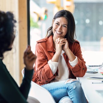 Heilpädagogen sind in vielen Bereichen tätig Zwei Frauen sitzen an einem Tisch in einem modernen Raum. Eine Frau mit braunen Haaren und einem orangefarbenen Mantel lächelt und schaut die andere an, die in einem grünen Oberteil gekleidet ist.