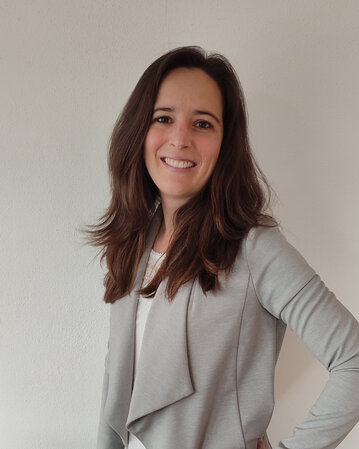 Eine Frau mit langen, braunen Haaren trägt einen grauen Blazer und lächelt. Der Hintergrund ist neutral und hell.