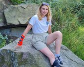 Eine Person sitzt auf einem großen Stein, trägt ein weißes T-Shirt und kurze Hosen. Sie hat eine rote Handbandage und eine Schiene am Bein. Im Hintergrund sind grüne Pflanzen sichtbar.