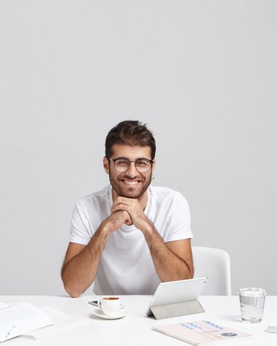 Ein junger Mann mit Brille sitzt an einem Tisch, lächelt und stützt sein Kinn auf die Hände. Vor ihm liegen Bücher, ein Notizbuch und eine Tasse. Ein Tablet steht auf dem Tisch.