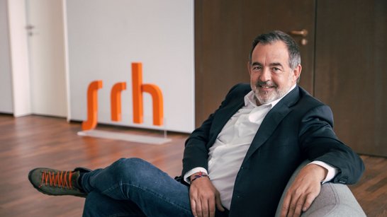 Ein Mann sitzt entspannt auf einem grauen Sofa, trägt einen schwarzen Blazer und Jeans, mit einem orangefarbenen Schriftzug "srh" im Hintergrund.