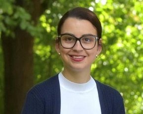 Eine Person mit Brille und einem blauen Cardigan lächelt vor einer grünen, bewaldeten Kulisse.