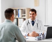 Ein Arzt in einem weißen Kittel sitzt an einem Tisch und spricht mit einem Patienten. Der Arzt hat ein Stethoskop um den Hals und lächelt, während der Patient ihm gegenüber sitzt.