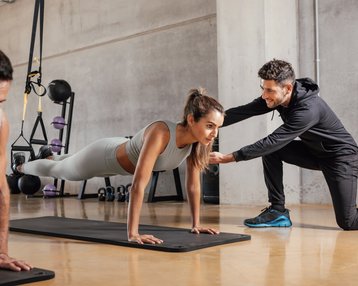Sportpsychologie Bachelor-Fernstudium Eine Frau führt Liegestütze auf einer Matte aus, während ein Trainer sie anleitet. Im Hintergrund sind weitere Sportler und Fitnessgeräte zu sehen.