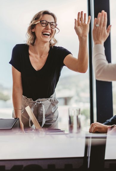 Eine Frau mit Brille steht an einem Tisch und gibt einer anderen Person ein High-Five. Beide lächeln, während sie in einem modernen Büro mit großen Fenstern sitzen.