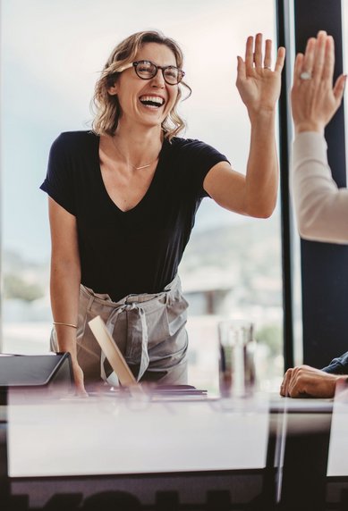 Eine Frau mit Brille steht an einem Tisch und gibt einer anderen Person ein High-Five. Beide lächeln, während sie in einem modernen Büro mit großen Fenstern sitzen.