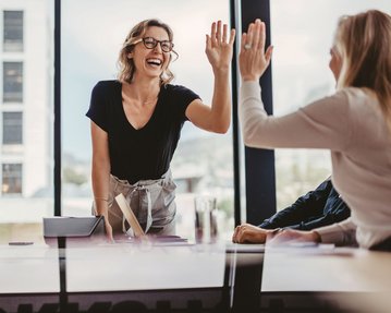 Change Management Eine Frau mit Brille steht an einem Tisch und gibt einer anderen Person ein High-Five. Beide lächeln, während sie in einem modernen Büro mit großen Fenstern sitzen.