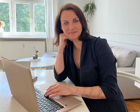 Eine Frau mit kurzen, braunen Haaren sitzt an einem Tisch und arbeitet an einem Laptop. Sie trägt ein schwarzes Oberteil und hat ihre Hand unter das Kinn gelegt, während sie in die Kamera schaut.