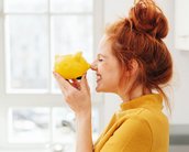 Eine Person mit roten Haaren hält ein gelbes Sparschwein vor ihr Gesicht und lächelt. Sie trägt ein gelbes Oberteil und steht in einer hellen, modernen Küche.