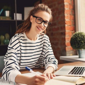 Eine junge Frau mit Brille sitzt an einem Tisch, schreibt in ein Notizbuch und lächelt. Auf dem Tisch liegen ein Laptop und einige Unterlagen. Im Hintergrund sind Pflanzen und Bücherregale zu sehen.