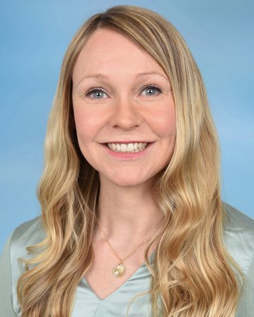 Eine lächelnde Frau mit langen, blonden Haaren trägt eine Halskette. Der Hintergrund ist hellblau.