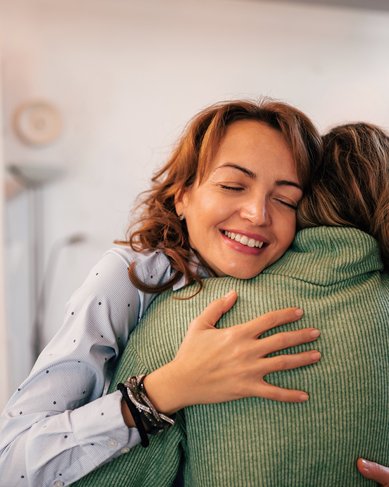 Zwei Frauen umarmen sich in einem modernen Raum. Eine Frau trägt ein hellblaues Hemd mit Punkten, während die andere einen grünen Pullover anhat. Beide lächeln und scheinen sich zu freuen.